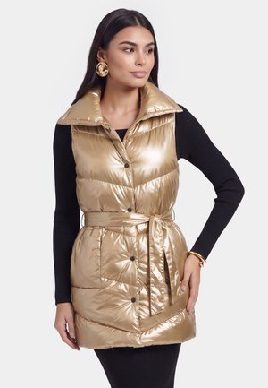 Frau trägt eine glänzende goldene Steppweste mit weitem Kragen und Gürtel über einem schwarzen Langarmshirt, ergänzt durch goldene Ohrringe und Armband.