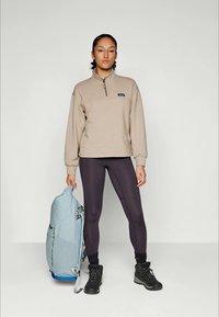 Beige fleece pullover met een halve rits en logo, gecombineerd met zwarte leggings en stevige zwarte laarzen. Lichtblauwe rugzak met verstelbare schouderbanden.
