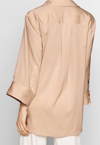 Beige silkkirtta, joka on rento malli, pitkähiha, ja napitus yksityiskohtana hihansuissa. Takana on poimutus, joka lisää laskostusta.