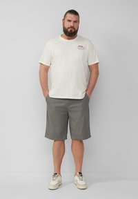T-shirt beige clair à manches courtes avec un motif rouge, associé à un short gris arrivant au genou et des baskets blanches avec des accents beiges.