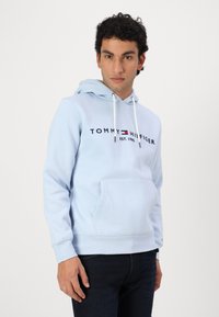 Ανοιχτό μπλε φούτερ με κορδόνια, μπροστινές τσέπες καγκουρό και εκτυπωμένο λογότυπο "TOMMY HILFIGER EST. 1985." Φτιαγμένο από μαλακό βαμβακερό μείγμα.