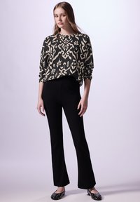 Maglione nero con motivo e design bianco, abbinato a pantaloni neri a palazzo e scarpe ballerine decorate. Modella in piedi contro uno sfondo chiaro.