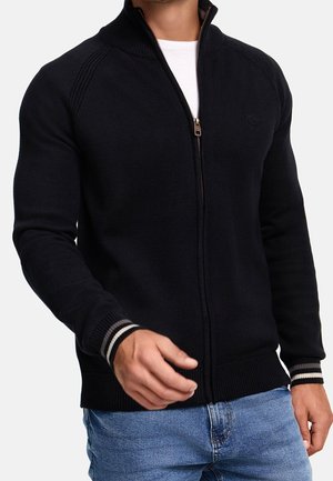 Homme portant un pull à fermeture éclair noir avec des poignets rayés sur une chemise blanche et un jean bleu, la fermeture éclair partiellement ouverte.