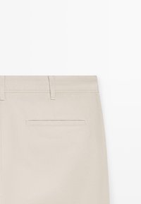 Pantalon beige avec poche passepoilée à l'arrière et passants de ceinture sur fond blanc.