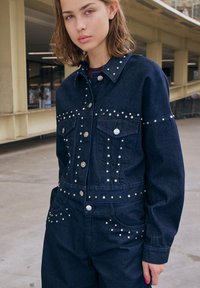 Denim kombinezon temno modre barve s srebrnimi žebljički ob ramenih, žepih na prsih in pasu. Ima kovinske gumbke in strukturirano prileganje.