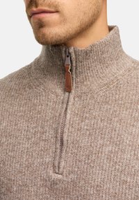 Lys brun, struktureret sweater med opretstående krave og halvlynlås; har en lædertrækklap og ribbede manchetter.