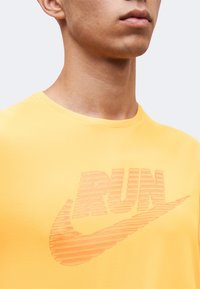 Κοντινό ενός ατόμου που φορά κίτρινο πουκάμισο με τη λέξη "RUN" και ένα λογότυπο swoosh σε ριγέ μοτίβο στο στήθος.