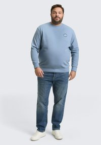 Man staat in een lichtblauwe sweatshirt, blauwe spijkerbroek en witte sneakers tegen een effen lichtgrijze achtergrond.