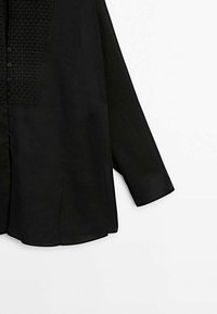 Chemise noire à manches longues avec boutons, panneau en tissu texturé en haut et un seul poignet sur la manche visible.