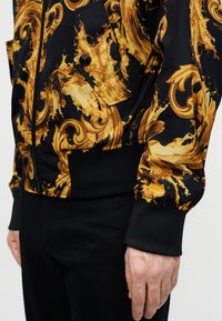 Μαύρο μπουφάν με χρυσούς στροβιλισμούς και πιτσιλιές σε μπαρόκ στυλ, με φερμουάρ μπροστά, τσεπάκι με κουμπί και ριμπ μανσέτες, φορεμένο με μαύρο παντελόνι.