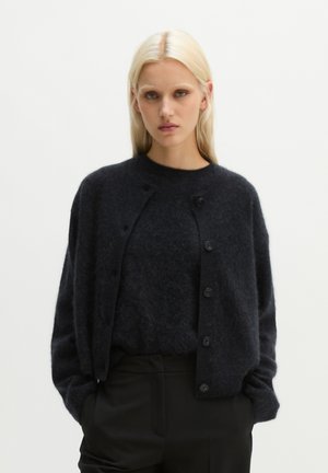 Donna bionda che indossa un cardigan nero peloso sopra un maglione abbinato, in piedi con le mani nelle tasche dei pantaloni neri su sfondo semplice.