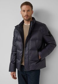 Pufasta jakna u tamnoplavoj boji, s visokim ovratnikom, dugmadi na pritiskanje i kvadratnim uzorkom. Uparena s bež džemperom i tamnim trapericama.