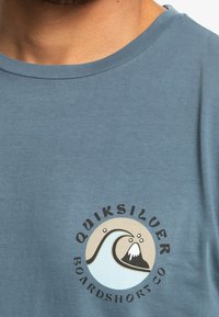 Tyrkysové bavlněné tričko s kulatým potiskem zobrazujícím vlnu a horský motiv. Černý text praví "Quiksilver Boardshort Co."