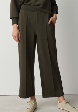 Personne portant un pantalon large vert olive avec des poches latérales et des baskets beiges, debout devant un fond clair uni.
