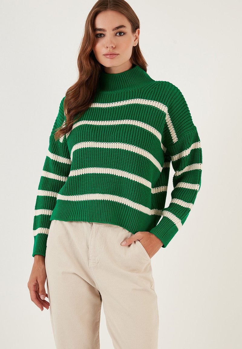 Maglione a righe verdi e bianche in maglia con collo alto e texture a coste, abbinato a pantaloni beige. Il design presenta maniche lunghe e una vestibilità rilassata.