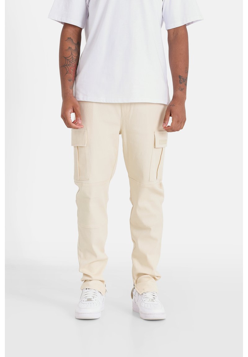 Beige cargobroek van dik materiaal met twee zijzakken, taps toelopende pijpen en zichtbare stiksels, gecombineerd met witte sneakers.