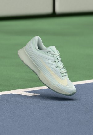 Light green Nike running shoe with beige swoosh hovering above a tennis court surface near white boundary lines.