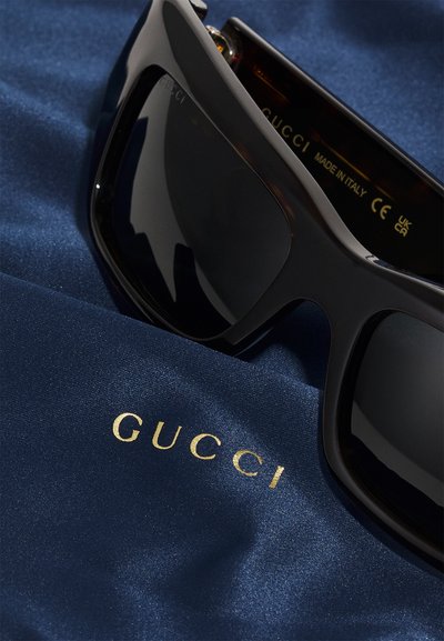 Occhiali da sole neri con lenti scure appoggiati su un tessuto liscio di colore blu navy. Il logo "GUCCI" in oro appare in modo prominente sul tessuto.