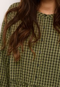 Camicia verde e nera a quadri con scollatura rotonda, chiusura frontale con bottoni e tessuto testurizzato, caratterizzata da un orlo arricciato.