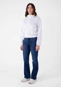 Giacca bianca con cappuccio, zip frontale e vita elastica, abbinata a jeans svasati blu e sneaker bianche. Tessuto liscio, design minimalista.