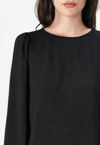 Svart langermet topp med rund halslinning og samlet skulderdetalj. Stoffet har en subtil tekstur og faller mykt.