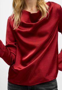 Röd satinblus med en tygkoll, långa ärmar och mjuk textur. Tyget faller följsamt och skapar en elegant silhuett.