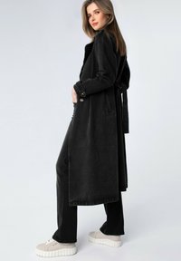 Lungo cappotto di denim nero con cintura da annodare, bottoni frontali e tasche laterali, abbinato a pantaloni neri e sneakers platform beige.