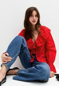 Blouse en coton rouge avec un col à volants et des broderies florales ; associée à un jean en denim bleu taille haute ; portée avec des ballerines noires.