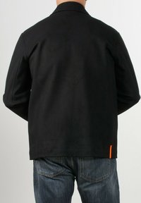 Giacca nera con colletto, tessuto testurizzato e un'accento arancione vicino al fondo. Indossata sopra jeans blu scuro, mostrando un look casual.