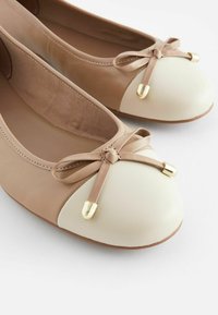 Ballet flats dengan bodi kulit beige, penutup jari putih, detail pita dengan aksen emas, dan lapisan interior yang berdimensi.