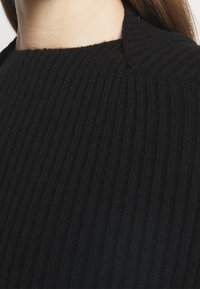 Schwarzer gerippter Strickpullover mit hohem Kragen und strukturiertem Oberflächen, der vertikale Rillen für zusätzlichen Detailreichtum und Dimension zeigt.