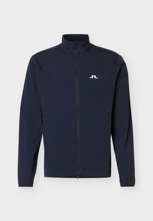 Lahka ženska jakna v mornarsko modri barvi z zadrgo po celotni sprednji strani, visokim ovratnikom, elastičnimi manšetami in majhnim belim logotipom na levi strani prsi.