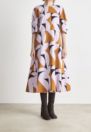Kvinde iført en mønstret midikjole med geometriske former i brun, lilla og sort, kombineret med sorte knæhøje støvler med hæl.