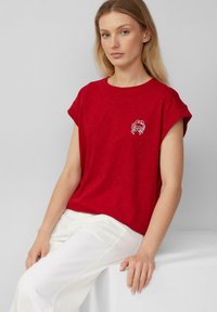 Vrouw met lang blond haar, gekleed in een rode kortlevenshirt met een klein wit krabontwerp, zittend op een witte blok, tegen een grijze achtergrond.