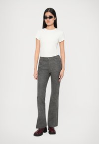 Γυναίκα με μακριά σκούρα μαλλιά, φορώντας μαύρα γυαλιά ηλίου, λευκή μπλούζα με κοντά μανίκια, γκρι υφασμάτινο παντελόνι και μπορντό πλατφόρμες, στέκεται μπροστά σε λευκό φόντο.