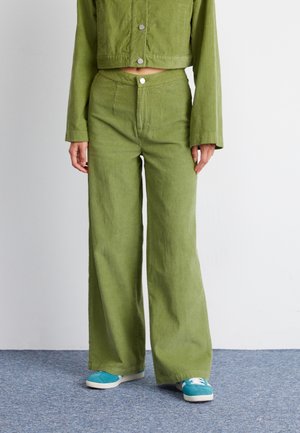Groene corduroy jas en bijpassende wijde broek met een gestructureerd oppervlak en zilveren drukknoopjes, gecombineerd met blauwe sneakers op een grijze vloer.