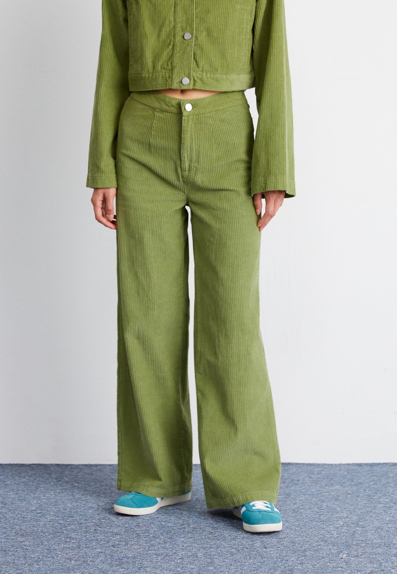 Groene corduroy jas en bijpassende wijde broek met een gestructureerd oppervlak en zilveren drukknoopjes, gecombineerd met blauwe sneakers op een grijze vloer.