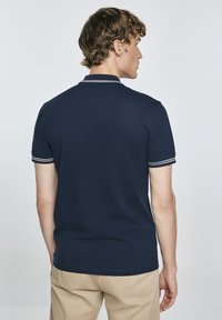 Tamsiai mėlyna polos marškinėliai trumpomis rankovėmis, su baltais akcentais ant apykaklės ir rankovių kraštų. Lygus audinys su standartiniu kirpimu.