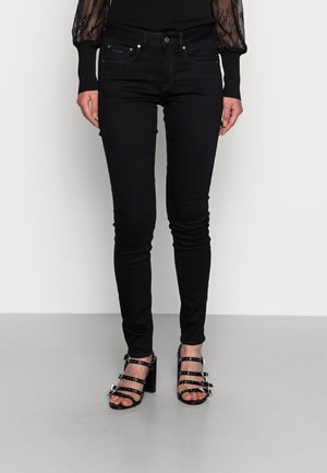 Frau trägt eng anliegende schwarze Jeans und schwarze High-Heel-Sandalen mit mehreren Schnallenriemen, steht vor einem einfarbigen hellen Hintergrund.