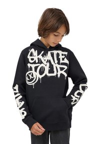 Ragazzo che indossa un hoodie nero con testo in stile graffiti bianco "SKATE TOUR" e un grafico di faccina sorridente, che guarda in basso con le mani in tasca.