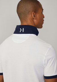 Bílá polo košile s námořnickým límcem a bílým písmem "H". Krátké rukávy s námořnickými akcenty, hladká textura a prodyšný materiál.