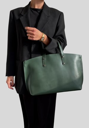 Borsa tote in pelle verde con due manici, texture liscia e dettagli in metallo. Il modello indossa un blazer nero e tiene la borsa all'altezza della spalla.