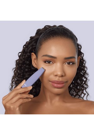 Femme aux cheveux bouclés utilisant un appareil de soin de la peau portable violet sur sa joue, sur un fond violet clair.