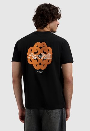 Man met krullend haar die een zwart T-shirt draagt met een oranje abstract ontwerp en de tekst "Pure Path" op de rug, gecombineerd met donkere spijkerbroek.