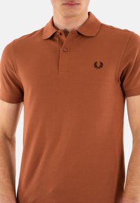 Bruine polo in een gestructureerde stof, met een kraag, drie knopen en een zwart embleem op de linkerkant van de borst. Korte mouwen.