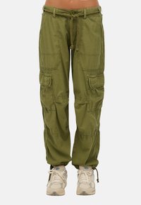 Olijfgroene cargo broek met meerdere voorzakken, een trekkoord in de taille en een gestructureerde stof. Gecombineerd met lichtgekleurde sportschoenen.