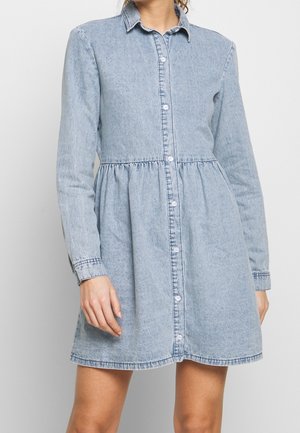 Vrouw die een lichtblauwe denim knoopdress met lange mouwen en een verzamelde taille draagt, staand tegen een effen achtergrond.