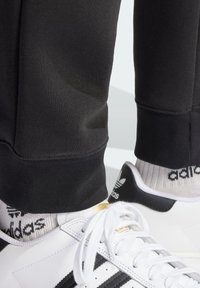 Svarta träningsbyxor med ribbade muddar, i kombination med vita sneakers med svarta ränder och gulddetaljer, samt mönstrade ankelsockor.