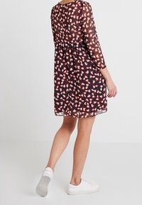 Pitkähihainen, polvipituinen mekko tummansinisellä värillä, jossa on värikäs abstrakti kuvio, varustettuna kerättyyn vyötäröön ja läpikuultavasta kankaasta. Yhdistettynä valkoisiin lenkkareihin.