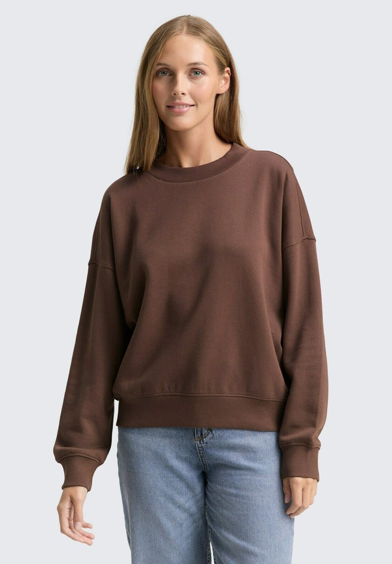 Brązowy bawełniany sweatshirt z okrągłym dekoltem, opuszczonymi ramionami i ściągaczami. W połączeniu z jasnoniebieskimi dżinsami. Gładka faktura.